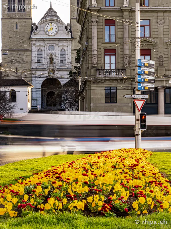View of the Hofkirche in Lucerne with a passing bus in the foreground and a flower bed