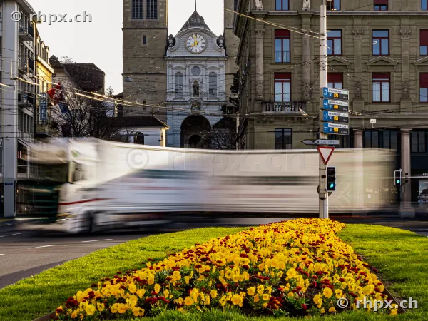 A truck drives past the Hofkirche in Lucerne with a long exposure, in the foreground a flower bed.