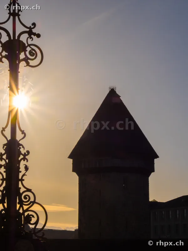 Silhouette of the Wasserturm in Lucerne against the low sun with an ornate lantern in the foreground.