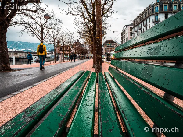Grüne Parkbank im Vordergrund auf der Uferpromenade in Luzern bei regnerischem Wetter