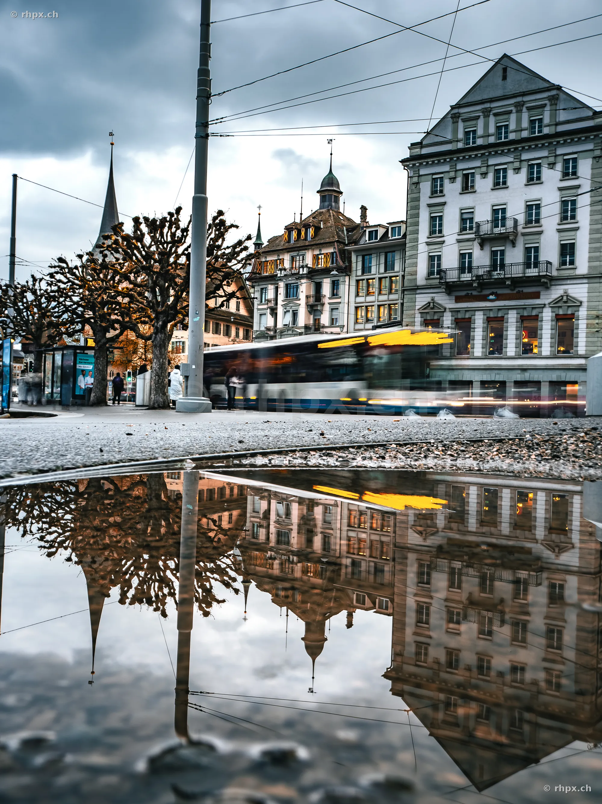 Spiegelung von Gebäuden und einem vorbeifahrenden Bus im Schweizerhofquai in einer Pfütze bei bewölktem Himmel.