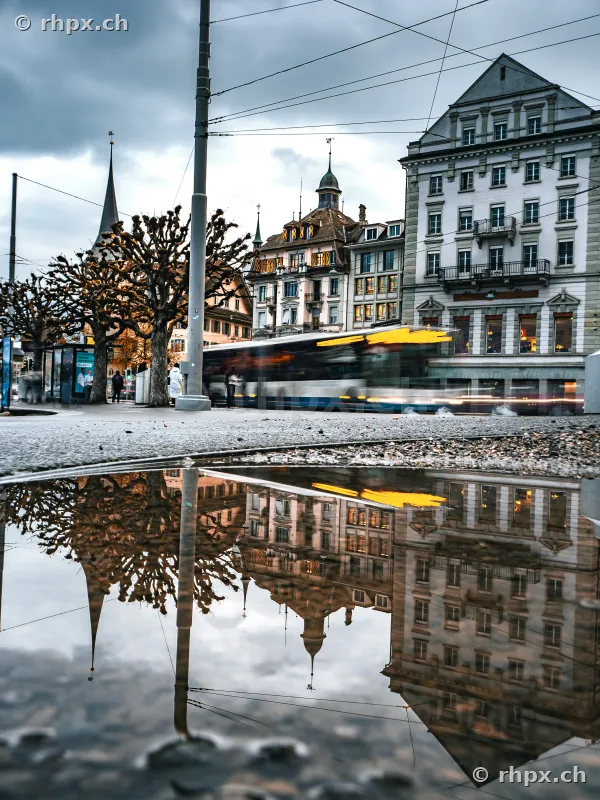 Spiegelung von Gebäuden und einem vorbeifahrenden Bus im Schweizerhofquai in einer Pfütze bei bewölktem Himmel.
