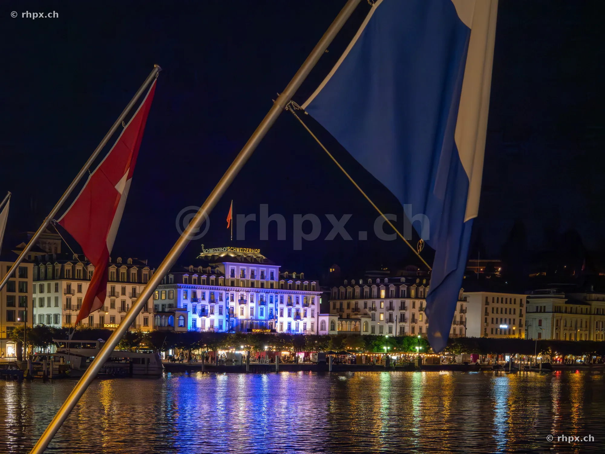 Blick über den Vierwaldstättersee auf das beleuchtete Hotel Schweizerhof in Luzern mit wehenden Flaggen im Vordergrund.
