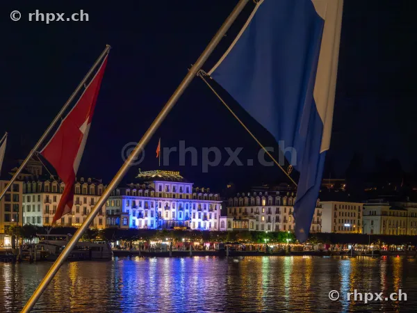 Blick über den Vierwaldstättersee auf das beleuchtete Hotel Schweizerhof in Luzern mit wehenden Flaggen im Vordergrund.