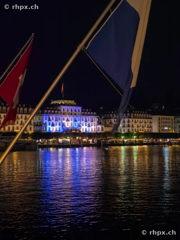Blick vom See auf das beleuchtete Hotel Schweizerhof in Luzern bei Nacht mit Flaggen im Vordergrund