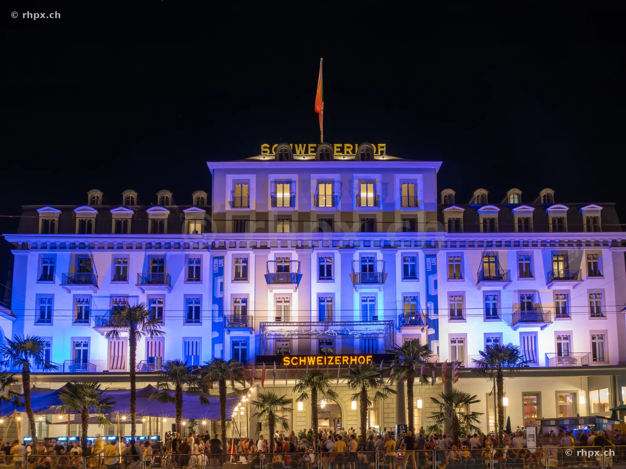 Fassade des Hotel Schweizerhof Luzern nachts blau beleuchtet mit Menschenmenge im Vordergrund