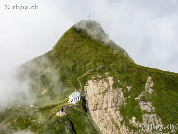 Small white chapel on a green mountain slope below the Pilatus summit with passing fog