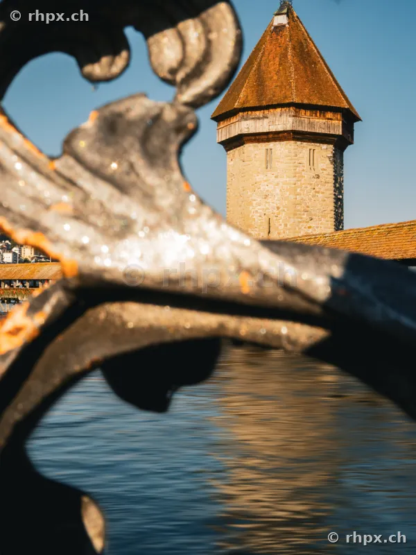 View of the Water Tower of the Chapel Bridge in Lucerne through an ornamental iron railing on the shore.