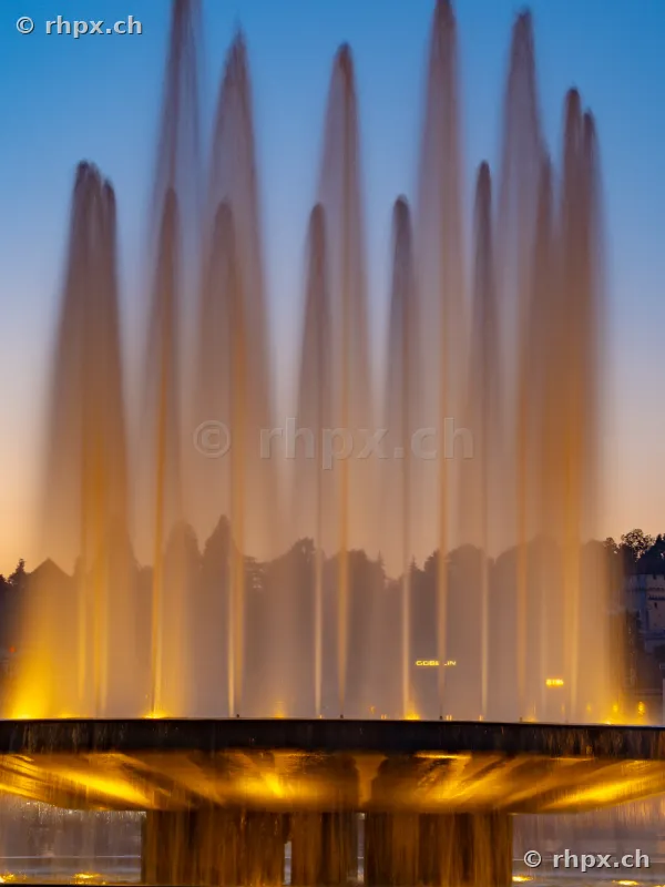 Illuminated fountain with tall water jets against a blue evening sky in Lucerne