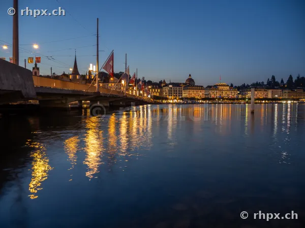 The Seebrücke bridge with flags and the illuminated old town of Lucerne reflecting in the water of Lake Lucerne at dusk.