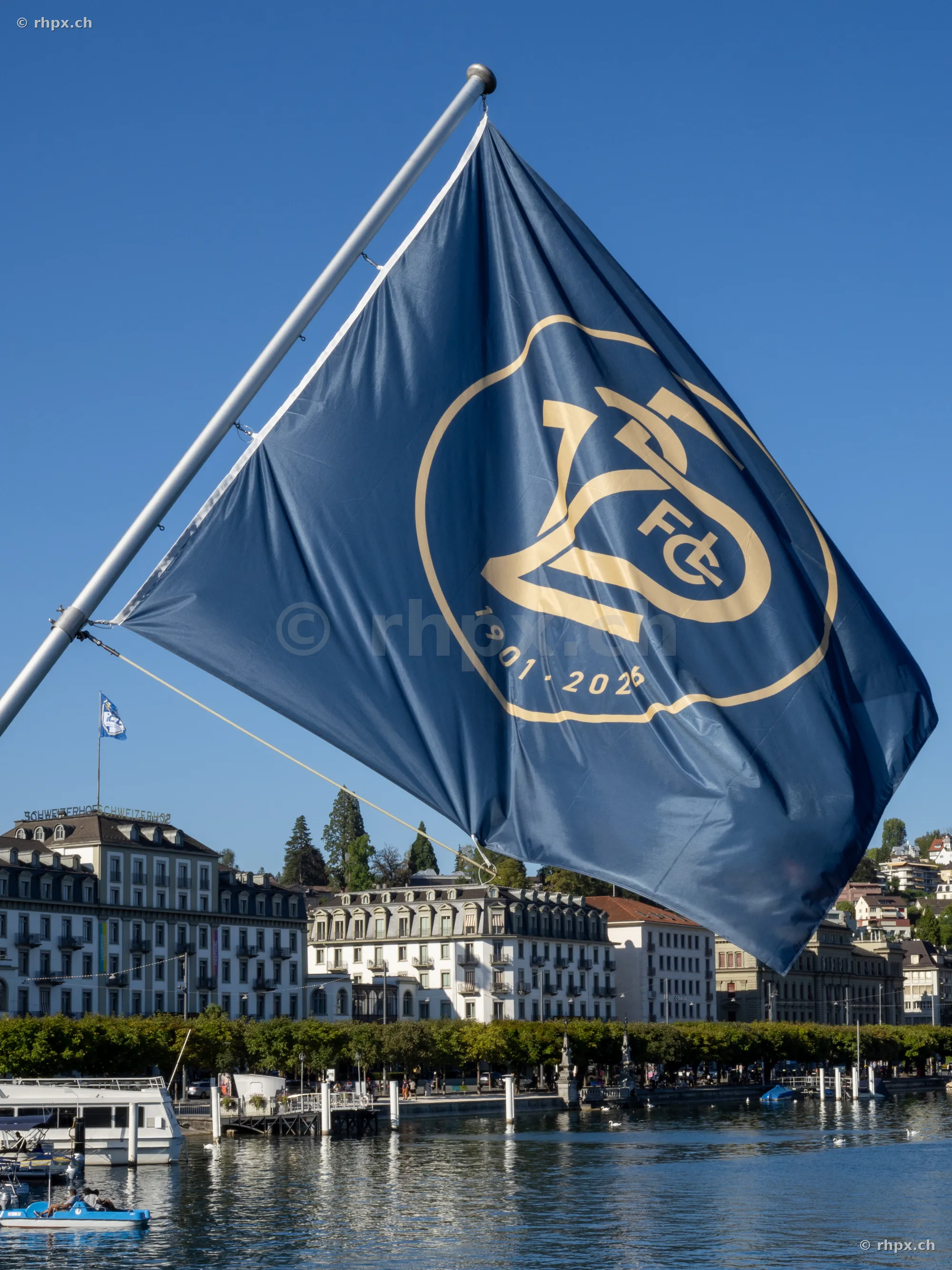 A dark blue anniversary flag of FC Luzern blowing in front of the Lucerne lakeshore scenery.