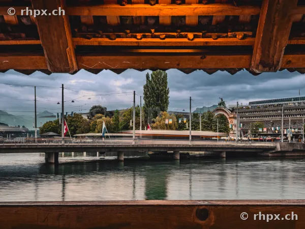 View through the timber framing of the Chapel Bridge towards the bridge and Lucerne railway station under a cloudy sky