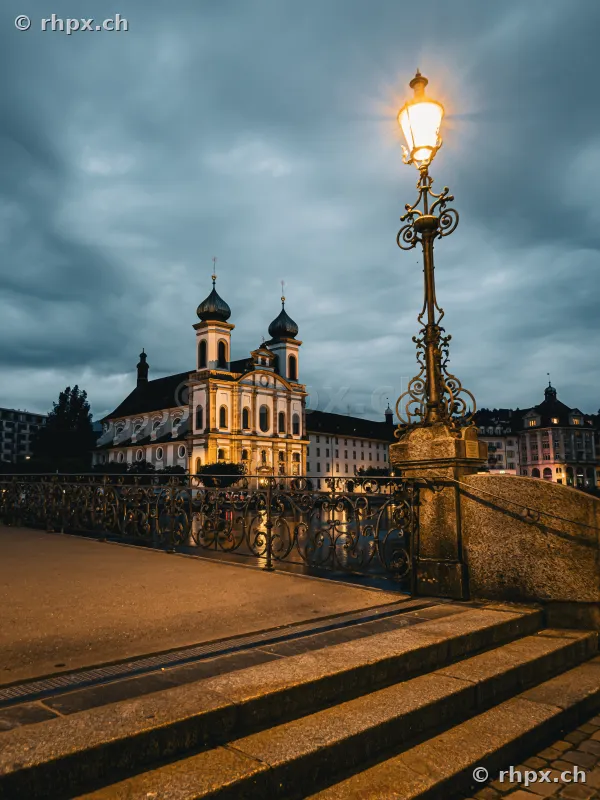 The illuminated Jesuit Church in Lucerne under a cloudy evening sky with a historic street lamp in the foreground.