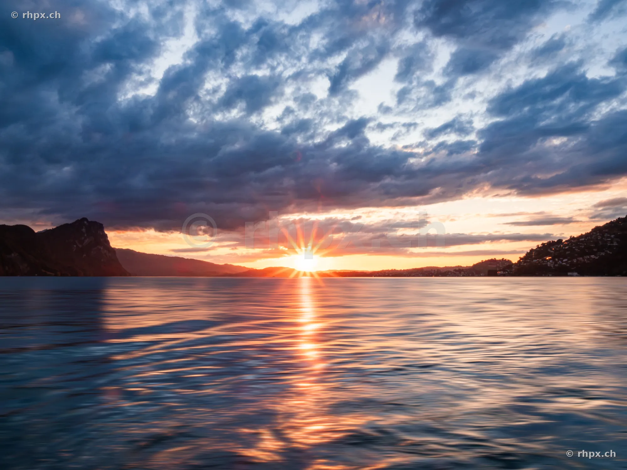Sun setting behind mountains over Lake Lucerne with cloudy sky
