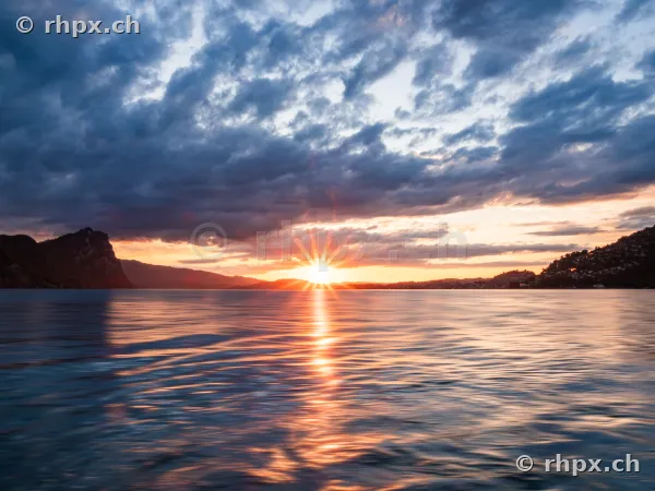 Sun setting behind mountains over Lake Lucerne with cloudy sky