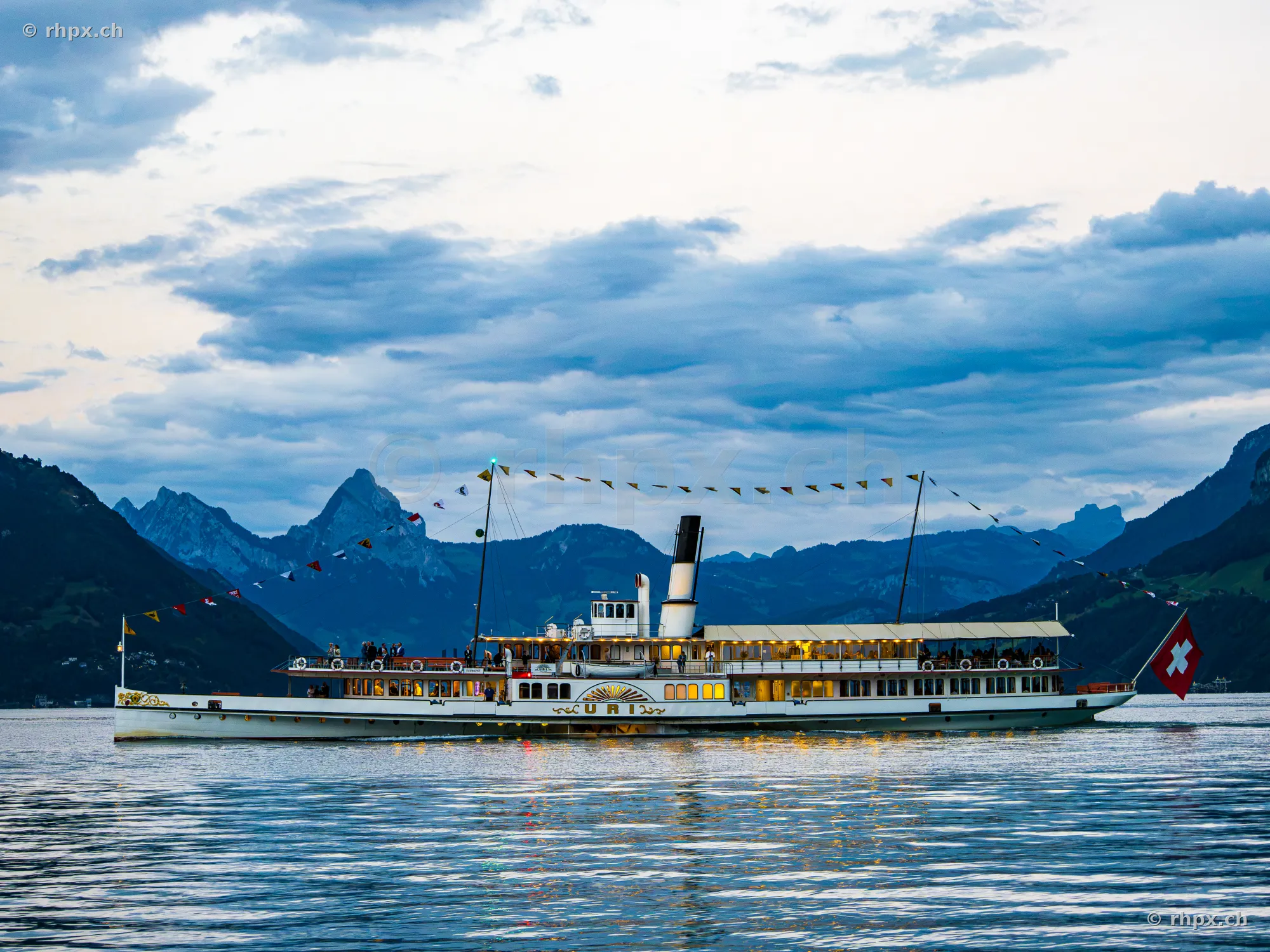 Das Raddampfschiff Uri fährt bei Abenddämmerung über den Vierwaldstättersee vor einer Bergkulisse