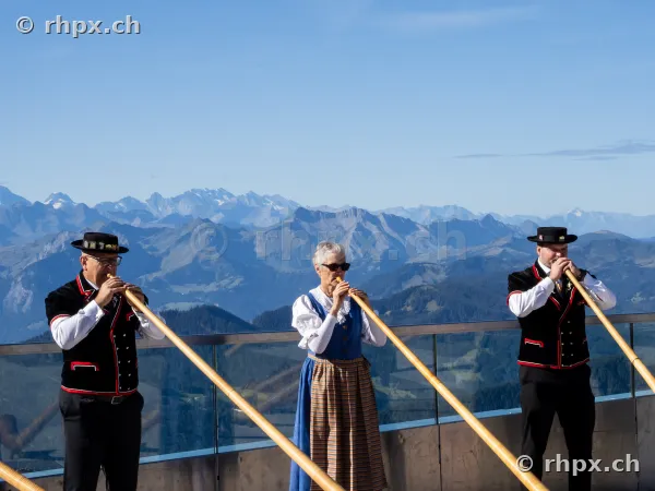 Two men and one woman in traditional Swiss costume playing alphorns on the observation deck of Mount Pilatus with an alpine panorama in the background.