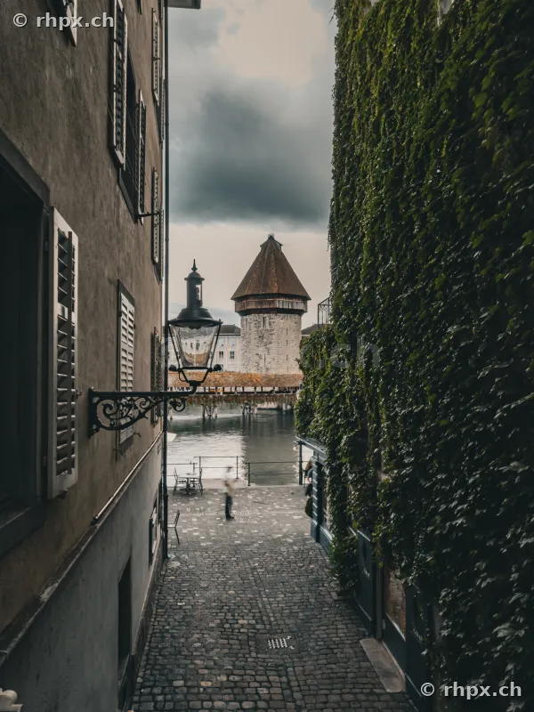 Blick durch eine enge Gasse auf den Wasserturm und die Kapellbrücke in Luzern bei bewölktem Himmel