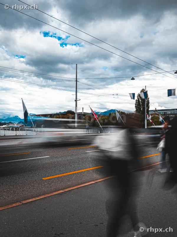 Blick auf die Seebrücke in Luzern mit fliessendem Verkehr und bewegten Passanten vor bewölktem Himmel.