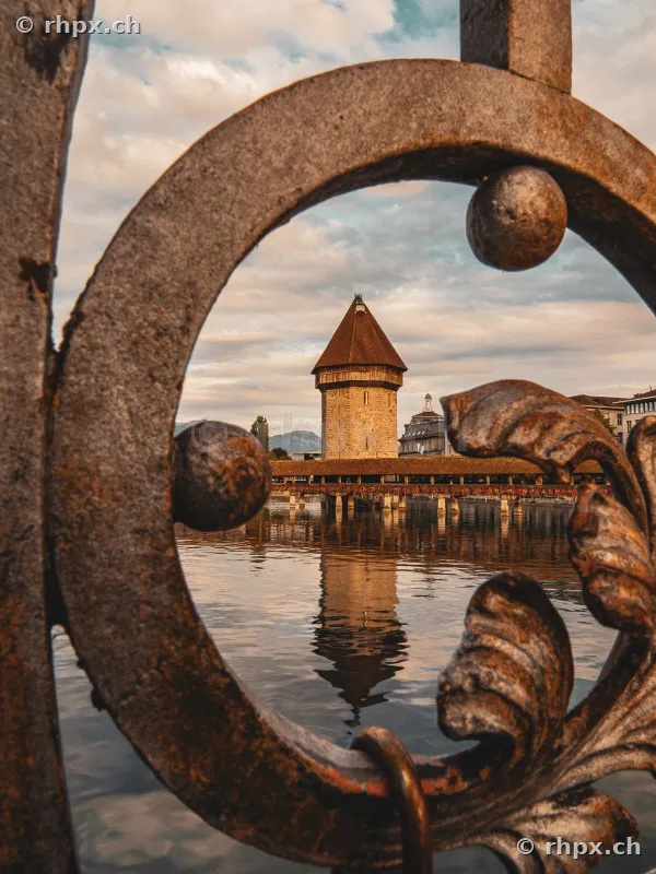Blick durch ein verziertes Metallgitter auf den Wasserturm und die Kapellbrücke in Luzern bei Abendlicht.