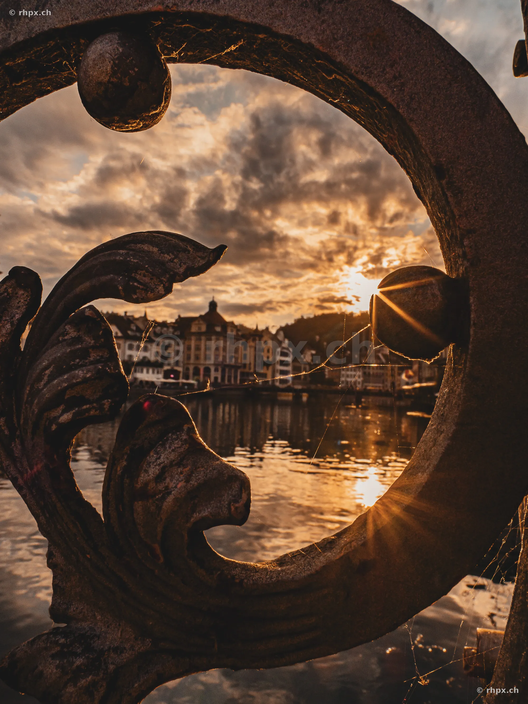 Blick durch ein verziertes Metallgeländer auf die Luzerner Altstadt und die Reuss bei Sonnenuntergang.