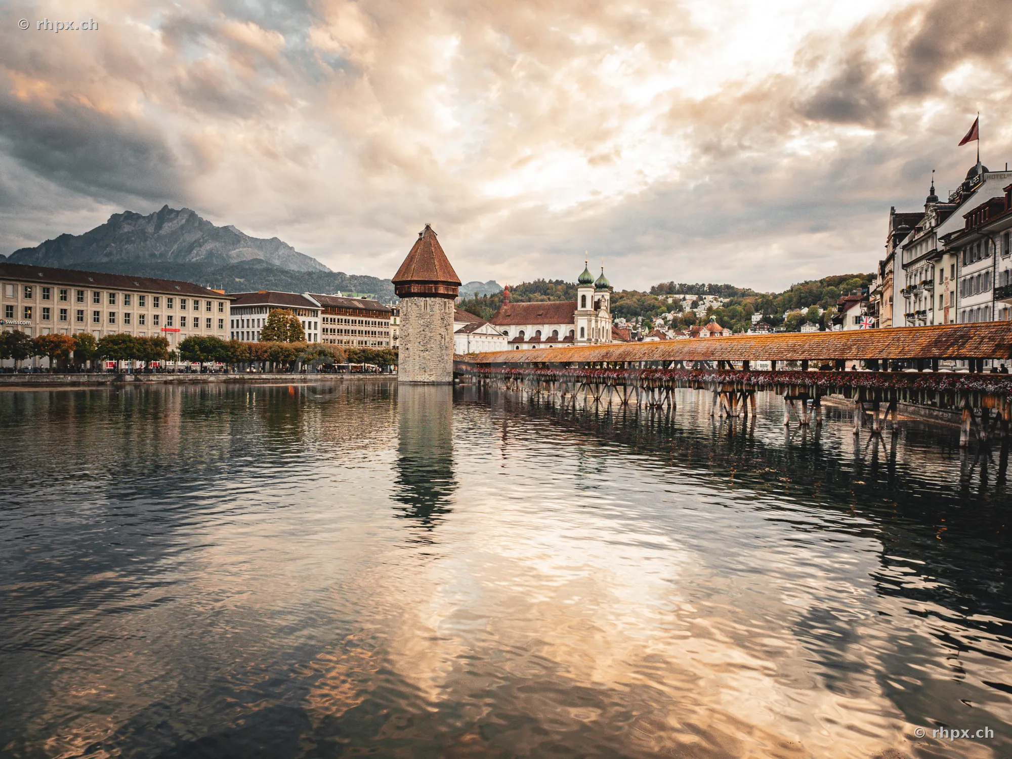 Blick auf die Kapellbrücke und den Wasserturm in Luzern mit dem Pilatus im Hintergrund bei bewölktem Abendhimmel.