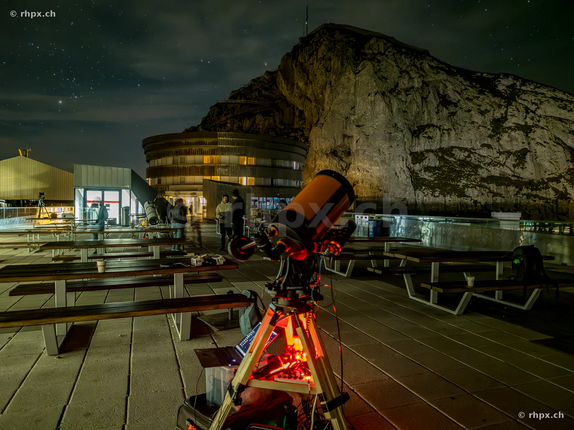 A telescope set up on the nighttime terrace in front of the Hotel Pilatus Kulm with the mountain massif in the background.