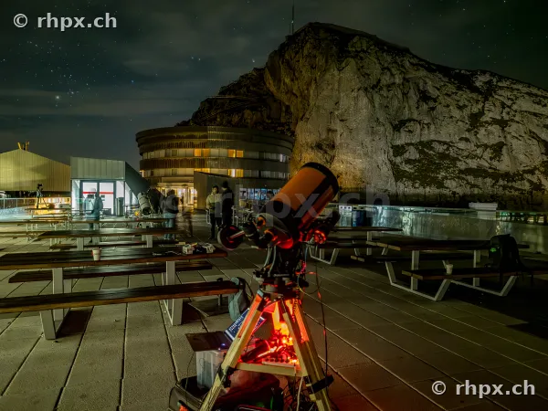 Ein aufgestelltes Teleskop auf der nächtlichen Terrasse vor dem Hotel Pilatus Kulm mit dem Bergmassiv im Hintergrund.