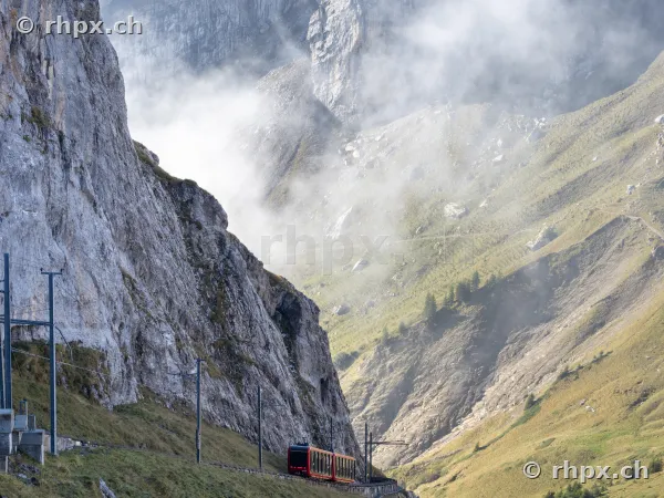 Rote Zahnradbahn fährt am steilen Felshang des Pilatus entlang bei nebligem Wetter.