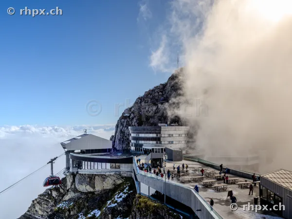 Blick auf das Hotel Pilatus Kulm und die ankommende Seilbahn bei teilweise bewölktem Himmel.