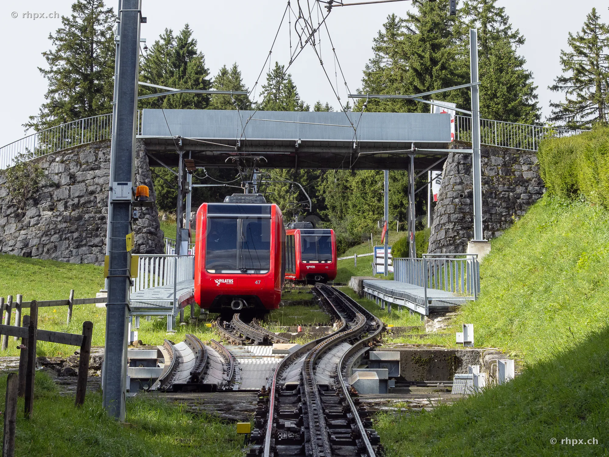 Two red Pilatus railway carriages traveling on tracks under a stone bridge