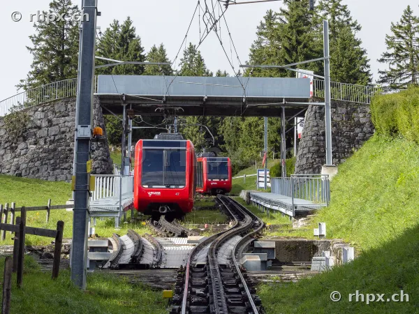 Two red Pilatus railway carriages traveling on tracks under a stone bridge
