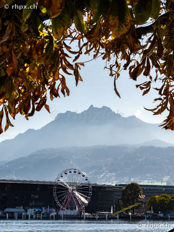 Riesenrad am Seeufer in Luzern mit dem Berg Pilatus im Hintergrund eingerahmt durch herbstliche Baumblätter