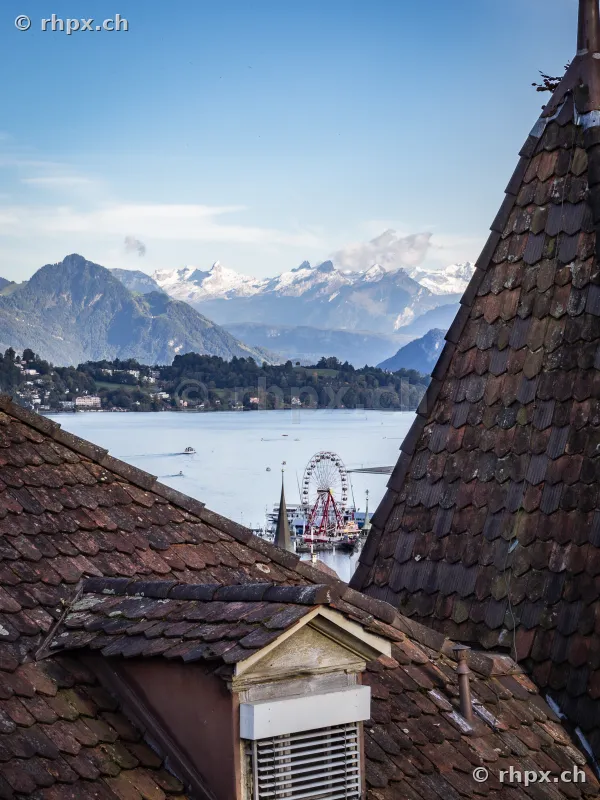 View between two tiled roofs looking towards Lake Lucerne with Ferris wheel and Alpine panorama