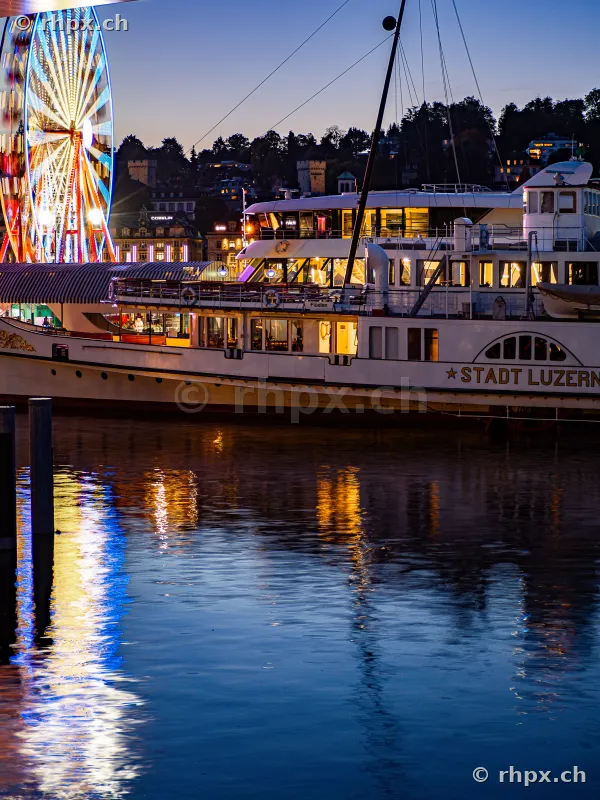 The paddle steamer Stadt Luzern docked at the shore in the evening with an illuminated Ferris wheel in the background.