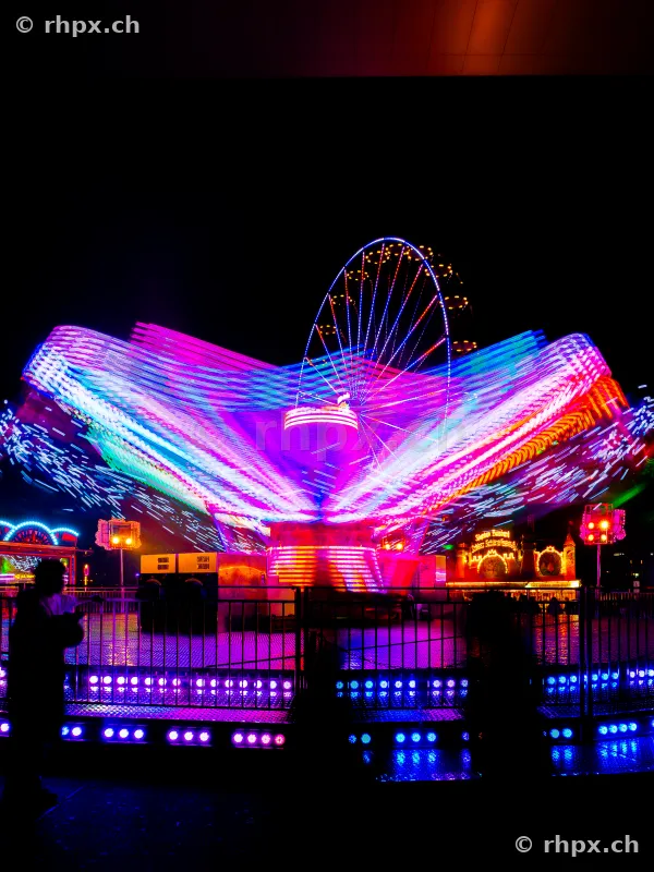 A swing ride in motion at night with light trails in front of a ferris wheel