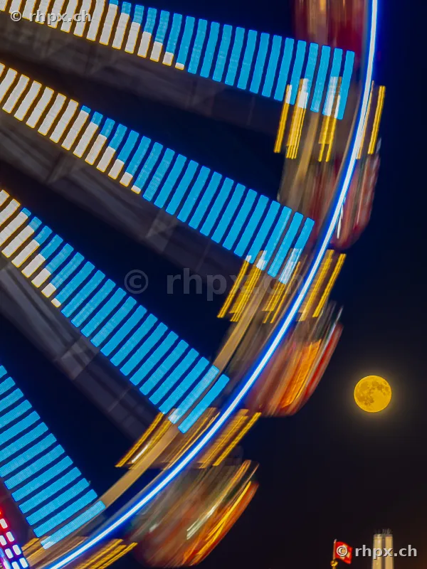 Long exposure of a Ferris wheel at night with a bright full moon and a Swiss flag in the background.