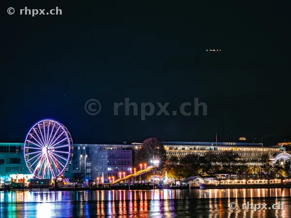 Illuminated Ferris wheel on the shore of Lake Lucerne at night, with the KKL Lucerne in the background.