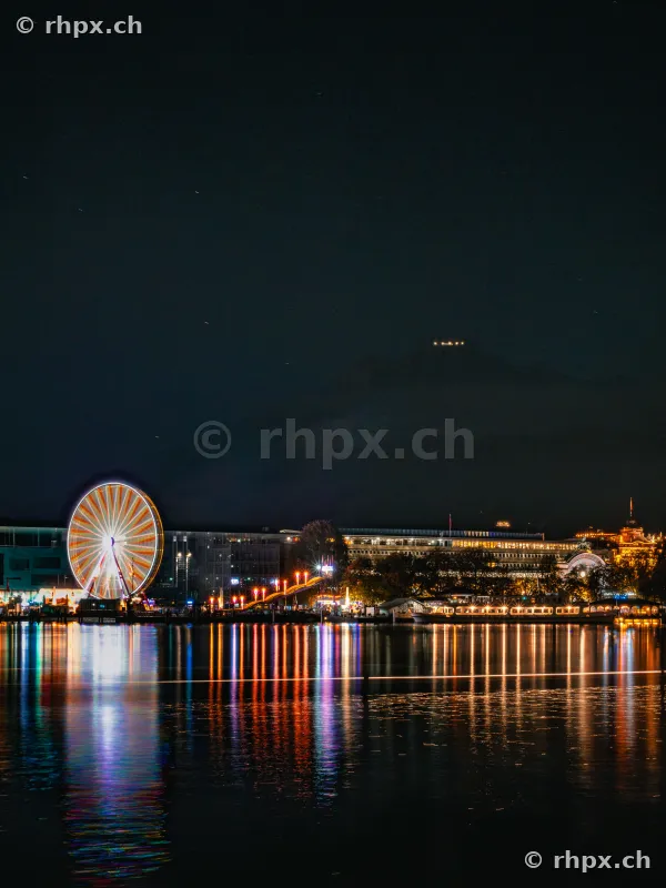 Illuminated Ferris wheel on the shore of Lake Lucerne at night with reflection in the water