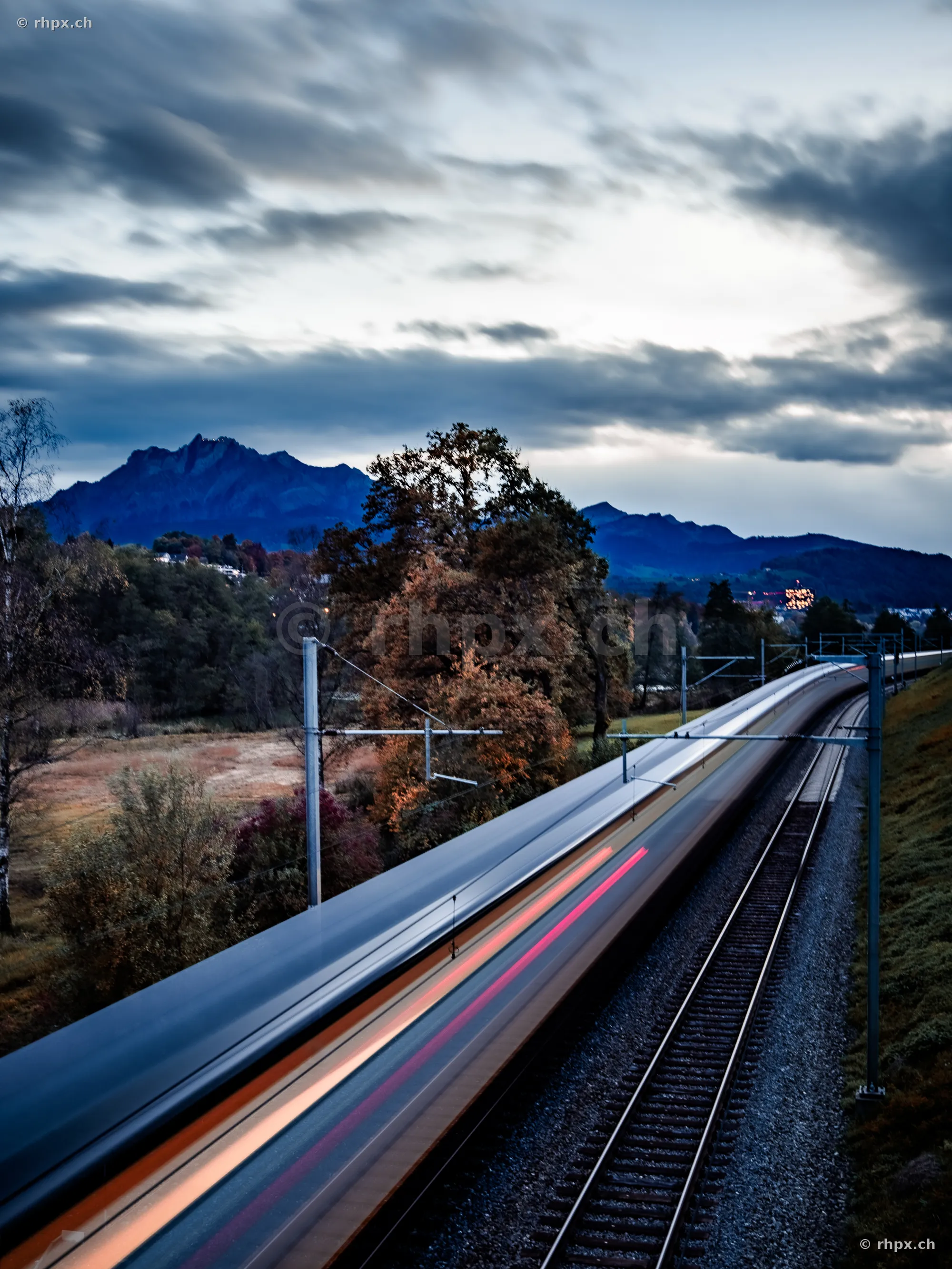 A motion-blurred train on the tracks in front of the Pilatus mountain scenery on a cloudy day.