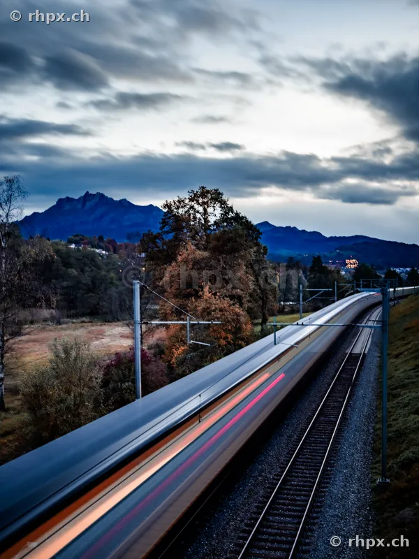 A motion-blurred train on the tracks in front of the Pilatus mountain scenery on a cloudy day.