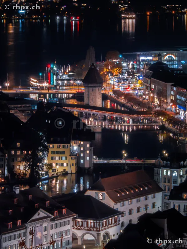 Blick von oben auf die beleuchtete Kapellbrücke in Luzern bei Nacht mit umliegenden Gebäuden am Flussufer.