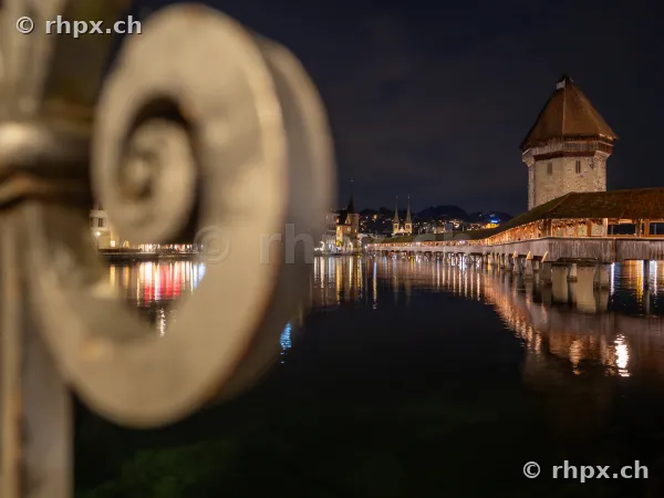 View of the illuminated Chapel Bridge in Lucerne at night, with a blurred ornamental railing in the foreground.