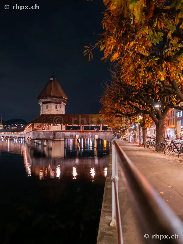The illuminated Chapel Bridge in Lucerne with autumnal trees along the riverbank at night