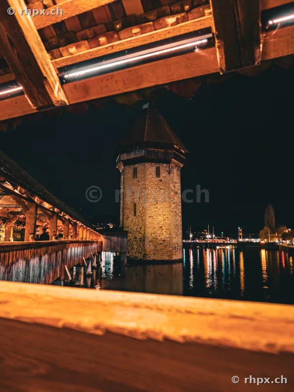 Blick durch den hölzernen Dachstuhl der Kapellbrücke auf den beleuchteten Wasserturm in Luzern bei Nacht.