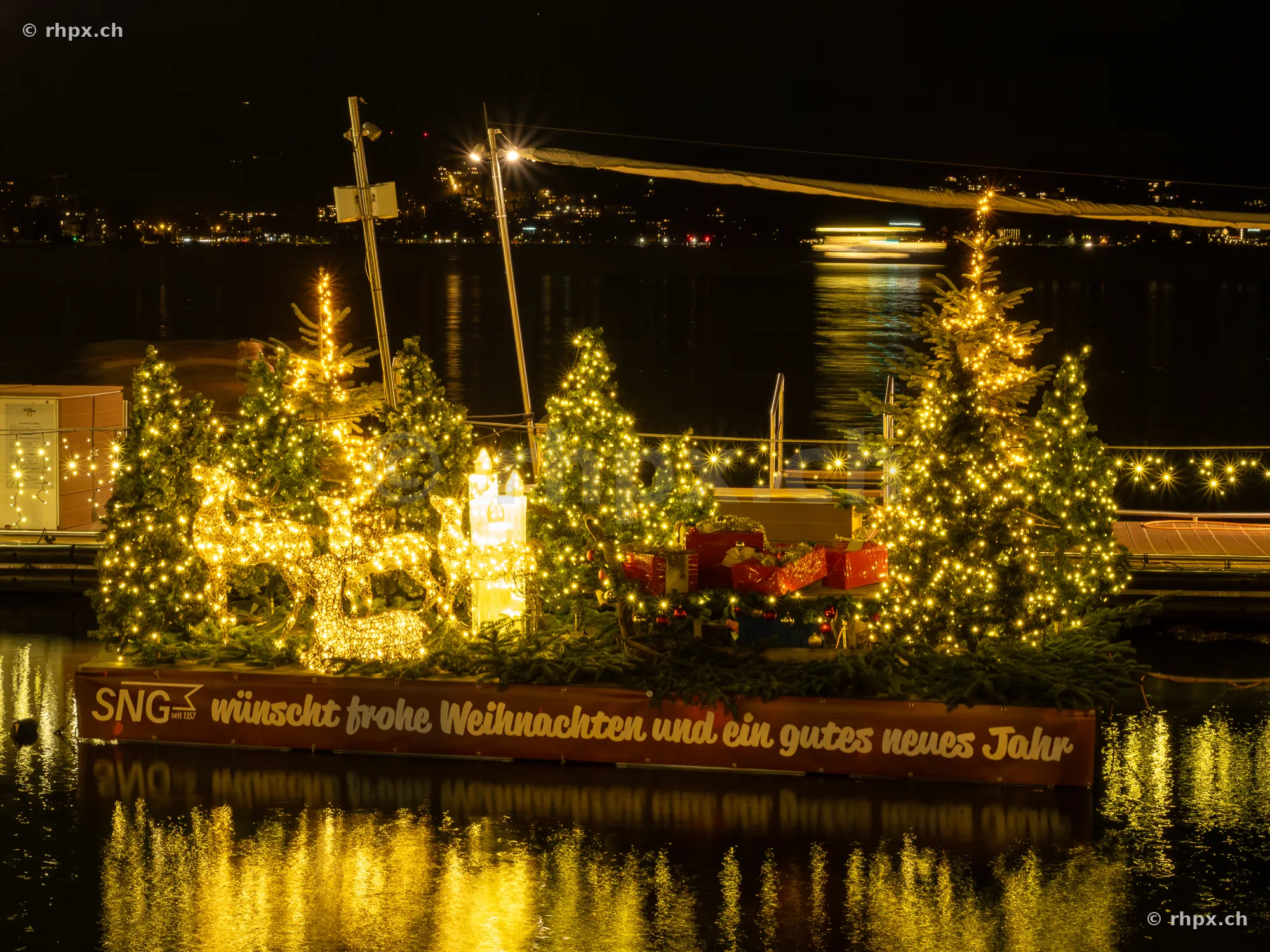 A Christmas-decorated raft with fairy lights, Christmas trees and reindeer figures floats on Lake Lucerne at night.