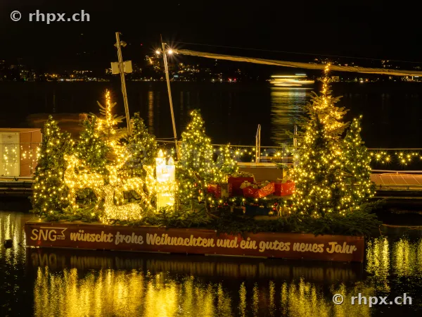 A Christmas-decorated raft with fairy lights, Christmas trees and reindeer figures floats on Lake Lucerne at night.