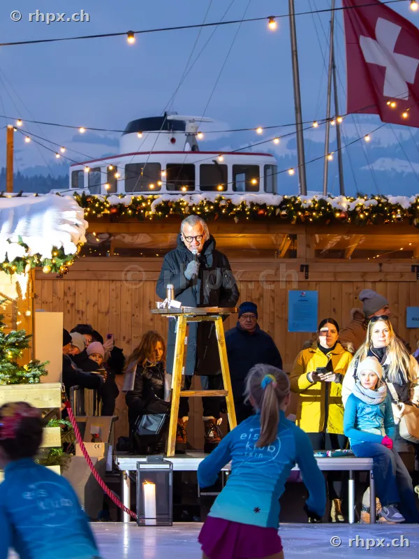 A person speaks at a lectern on an ice rink in Lucerne, with a boat and a Swiss flag visible in the background at dusk.