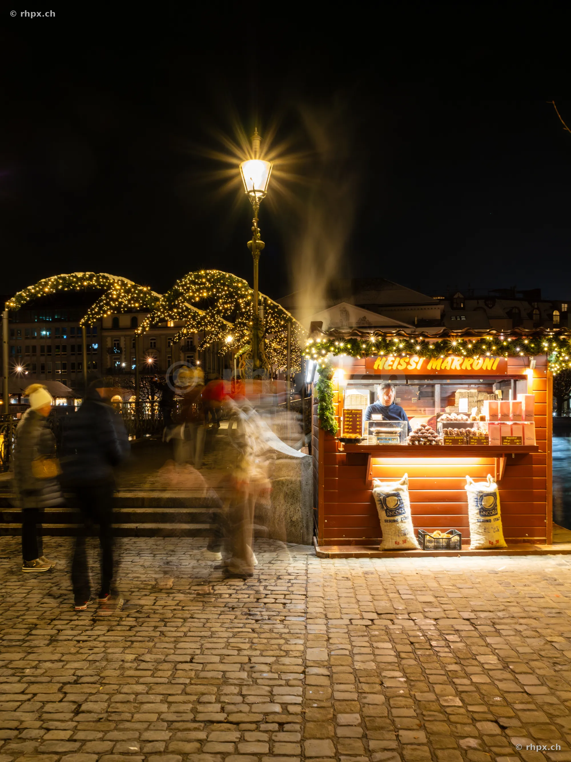 Ein beleuchteter Marroni-Verkaufsstand bei Nacht mit Passanten auf dem Kopfsteinpflaster in Luzern.