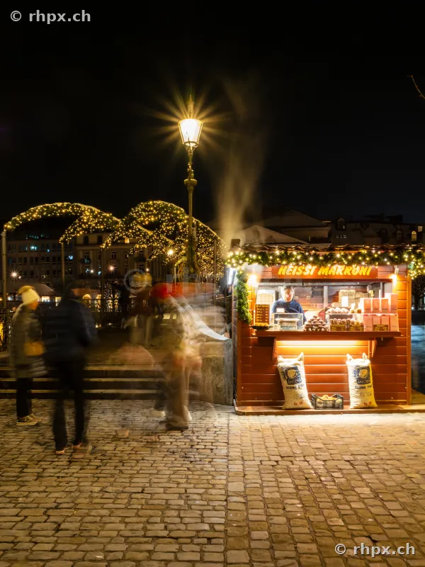 An illuminated chestnut vendor stand at night with pedestrians on cobblestones in Lucerne.